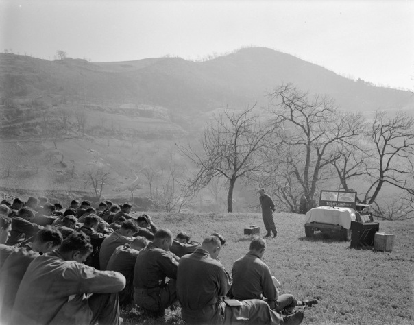 "One of many Easter services held on Appenine mountainsides by the Tenth Mountain Division April 1, 1945; conducted by Caplain William H. Bell for the 605th Artillery Battalion at Rocca Pitigliano. A large group of soldiers sit in a grassy open field with heads bowed. Before them stands the chaplain with a box beside him, a jeep marked beneath the windshield with 'Chaplain' in between two crosses, and a portable pump organ."