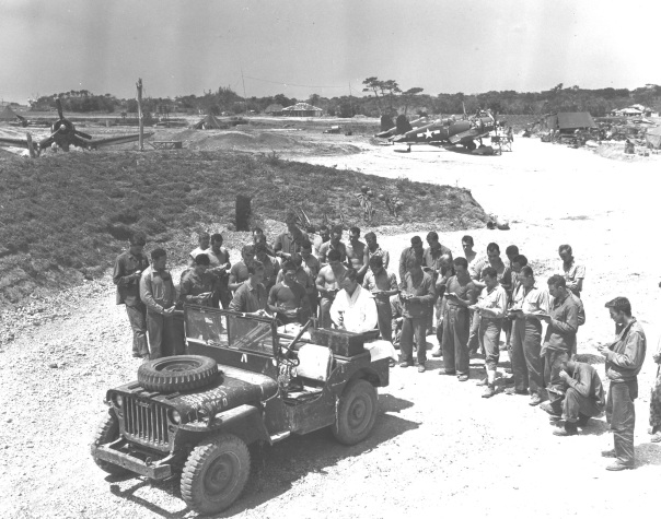 "Chaplain Joseph J. Garrity conducts the first Catholic services on Yontan airfield, Okinawa, 16 April 1945. The Hell's Belles fighter squadron of MGen Francis P. Mulcahy"