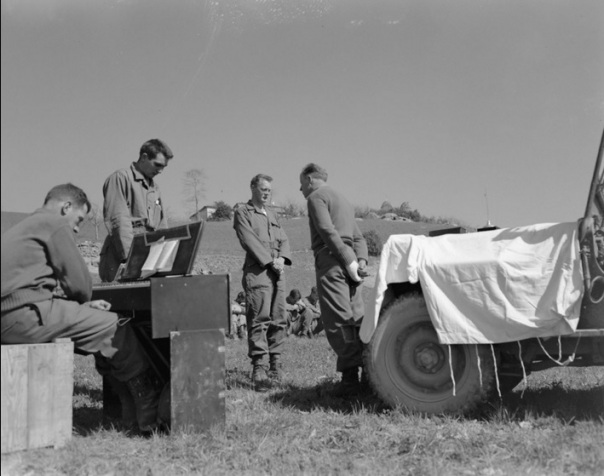 "April 1, 1945. Protestant Easter Service in Appennines, Italy background are trees and buildings.; Members of the Tenth Mountain Division, 605th Artillery Battalion, attend a Protestant Easter religious service at Rocca Pitigliano, Italy, conducted by Chaplain William H. Bell. In the foreground, four men bow their heads together. Corporal Ralph Squires sits at a portable organ and two soldiers face the Chaplain who stands in front of his jeep draped with a white cloth in use as an altar for a small crucifix."