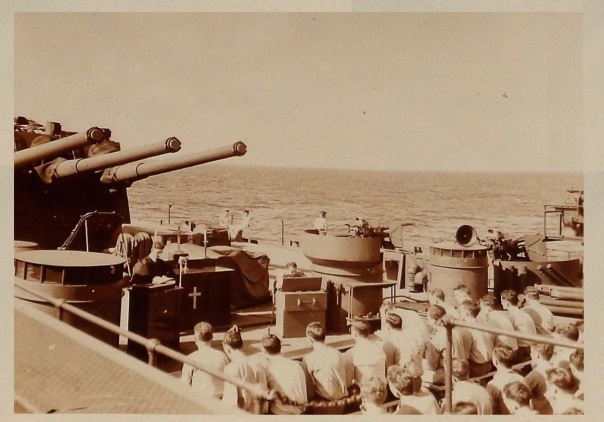 1940s, chaplain leads worship on the deck of ship in the shadow of the guns.
